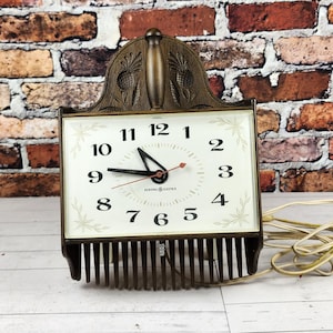 May include: Vintage General Electric electric clock with a square face and a decorative bronze-colored metal frame. The clock has a white face with black numbers and hands, and a red second hand. The clock is plugged in.