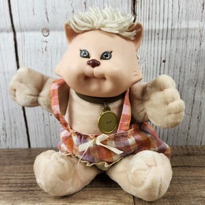 May include: A vintage plush cat doll with a light tan face, light blue eyes, and a small brown nose. The doll wears a plaid dress with suspenders and a gold-colored pendant. The doll has light blonde hair and is sitting.