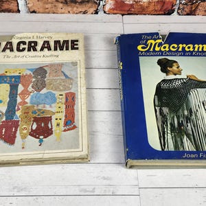 May include: Two vintage books on macrame. The book on the left is titled "Macrame: The Art of Creative Knotting" by Virginia I. Harvey. The book on the right is titled "The Art of Macrame: Modern Design in Knotting" by Joan Fisher. Both books are on a white wooden surface.