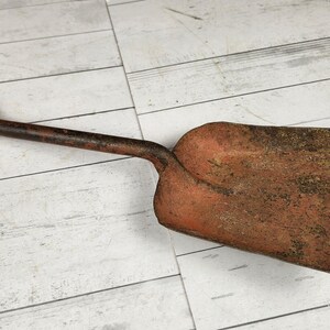 May include: An antique, rusty shovel with a wooden handle and a D-shaped grip. The shovel's blade is a weathered orange-brown color, and the handle is a dark brown. The tool is designed for digging and gardening.