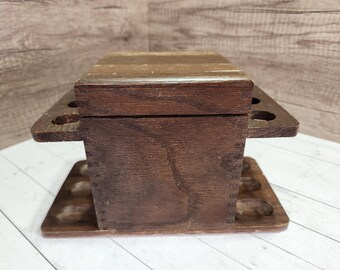 May include: A vintage wooden pipe stand and storage box. The box is rectangular with a hinged lid and a dark brown finish. It features multiple circular holes for holding pipes and a lower tray for accessories. The wood grain is visible.