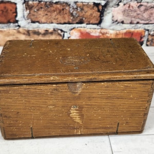 May include: A rectangular wooden box with a hinged lid. The box is made of light brown wood with visible grain and some wear. The box is closed and sits on a white wooden surface.