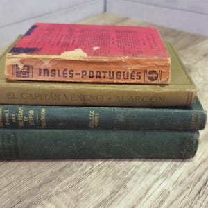 May include: A stack of four vintage books with aged covers. The top book is red with the title "INGLÊS-PORTUGUÊS". Below it is a green book with the title "EL CAPITÁN VENENO - ALARCÓN". The bottom two books are dark green.