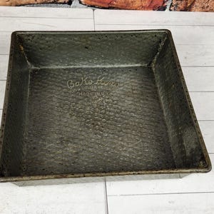 May include: A square metal baking pan with a dark, textured finish. The interior base features an embossed logo that reads Bakewell and includes the text Made in USA. The pan has straight, vertical sides and a flat bottom surface for baking.