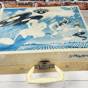 May include: Vintage children's record player case with a blue and white illustration on the lid. The artwork depicts musical notes, records, and cartoon figures. The case has a cream-colored handle and a metal clasp. The text "Playtime" is visible.