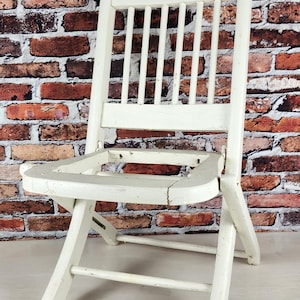 May include: A white wooden folding chair with a slatted backrest and a missing seat panel. The chair features a distressed paint finish with visible wear on the frame and legs, showcasing a simple construction with four legs and a horizontal support.