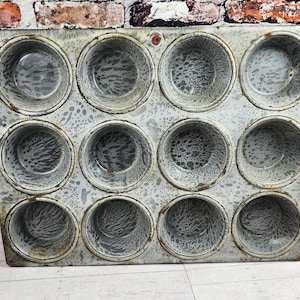 May include: Vintage metal muffin tin with twelve circular molds. The tin has a distressed, speckled gray finish, showing signs of wear. The background features a rustic brick wall and a wooden surface, suggesting a farmhouse aesthetic.