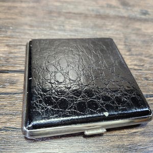 May include: A black leather cigarette case with a silver metal frame. The case has a textured surface and a worn look.