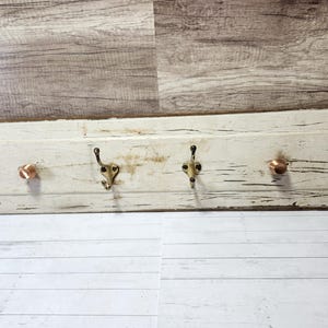 May include: White wooden coat rack with three hooks and two copper knobs. The rack has a distressed finish and is perfect for hanging coats, hats, or scarves.