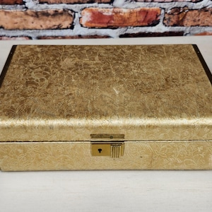 May include: A rectangular jewelry box with a textured gold finish and a metallic gold-toned trim. The box features a hinged lid and a central gold-colored latch mechanism with a keyhole on the front panel, presenting a uniform and structured design.