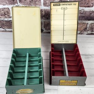 May include: Two vintage metal compartment files. One is teal with a cream-colored lid, the other is red with a cream-colored lid. The red file has a paper insert that reads "Compartment File" and has a measurement scale.