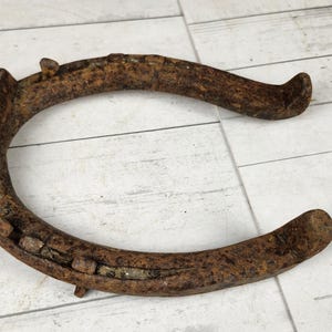 May include: A rusty, antique horseshoe made of iron. The horseshoe is curved with rounded ends and several nail holes along the bottom. The metal has a weathered, brown appearance, suggesting age and use. It rests on a light-colored, wooden surface.