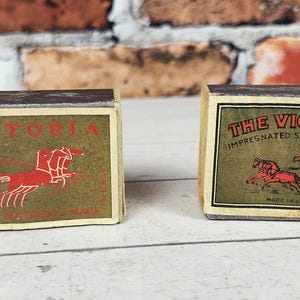 May include: Two vintage matchboxes with the text "VICTORIA" and "THE VICTORIA IMPREGNATED SAFETY MATCHES". The boxes have a cream-colored background with red and green graphics of horses and a chariot. Made in the USA.
