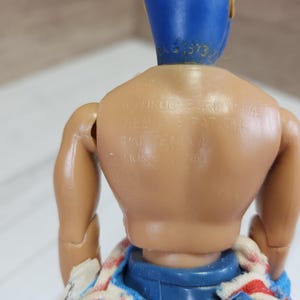 May include: A plastic action figure with a blue and yellow costume. The figure has a tan body and is wearing a blue and white striped shirt. The figure has the text "SHOO-FLY" printed on its back.