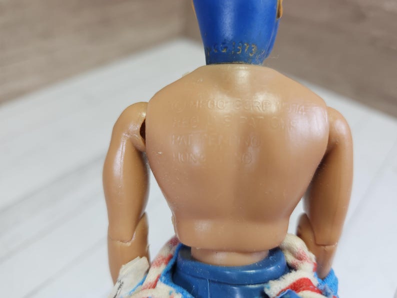 May include: A plastic action figure with a blue and yellow costume. The figure has a tan body and is wearing a blue and white striped shirt. The figure has the text "SHOO-FLY" printed on its back.