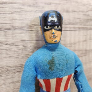 May include: A vintage action figure of Captain America wearing a blue and red costume with a blue and black mask. The figure has a faded paint job and is missing some of its original details.