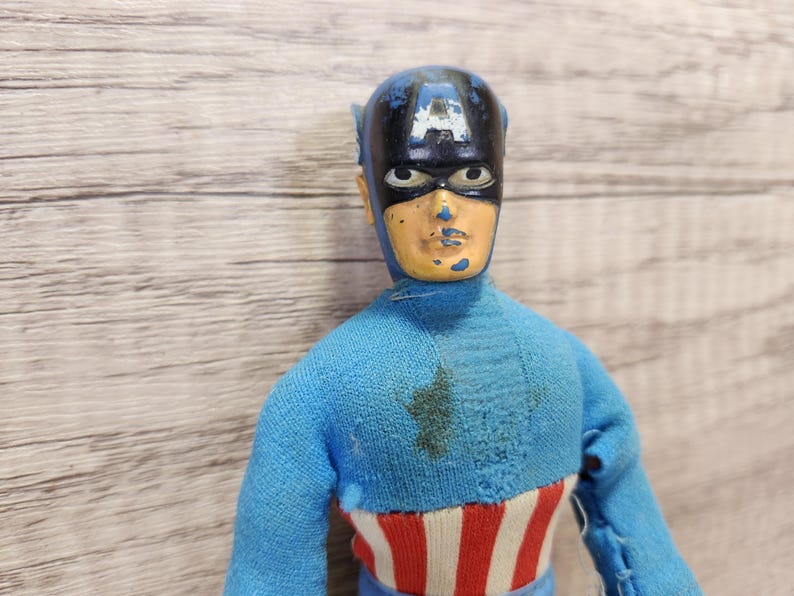 May include: A vintage action figure of Captain America wearing a blue and red costume with a blue and black mask. The figure has a faded paint job and is missing some of its original details.