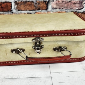 May include: A small, vintage-style suitcase with a cream-colored exterior and red trim. The suitcase features a metal clasp and handle, and is likely made of a durable material. The design is reminiscent of classic travel luggage.