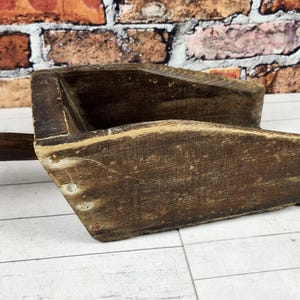 May include: A vintage wooden scoop with a long handle. The scoop is made of dark brown wood and has a rectangular shape with a deep bowl. The handle is rounded and attached to the side of the scoop. The wood has a weathered appearance.