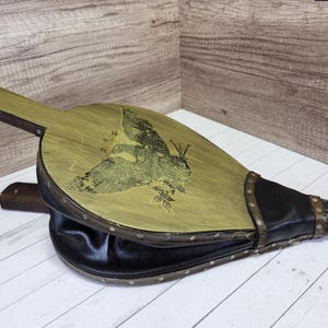May include: A vintage-style bellows with a green wooden panel and a black leather bellows. The wooden panel has a decorative design of a bird with a floral motif.