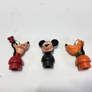 May include: Three plastic figurines of Disney characters: Goofy, Mickey Mouse, and Pluto. Goofy is wearing a red hat and red and white outfit. Mickey Mouse is wearing a black and white outfit. Pluto is wearing an orange and green outfit.