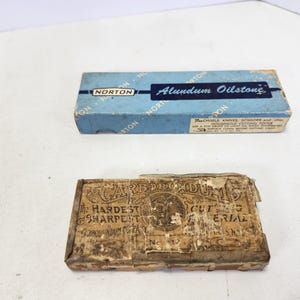May include: Two boxes of sharpening stones. The top box is blue and says "Norton Alundum Oilstone" and the bottom box is brown and says "Carborundum The Hardest Sharpening Material".