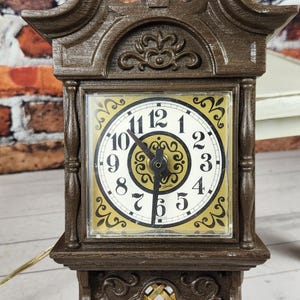 May include: A brown wall clock with a decorative design. The clock face has black hands and white numbers. The clock is rectangular with a decorative top and bottom. The clock is powered by a cord.