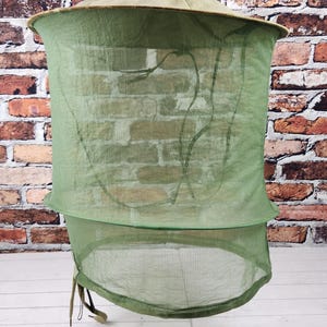 May include: A khaki-colored hat with a green mesh net attached. The net is designed to protect the face and neck from insects. The hat is sitting on a white surface in front of a brick wall.