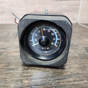 May include: A black and silver round gauge with a white face and black numbers. The gauge reads "RPM" and has a needle pointing to 40. The gauge is mounted in a black plastic housing.