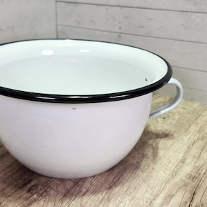 May include: White enamelware bowl with a black rim and handle. The bowl has a rounded shape and sits on a wooden surface. The interior of the bowl is white, and the rim is black. A classic kitchenware item.