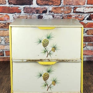 May include: A vintage two-drawer storage cabinet with a cream-colored finish and yellow trim. Each drawer features a pine cone and branch design. The cabinet is set against a brick wall and wooden surface.