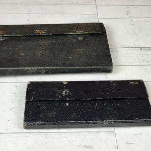 May include: Two vintage black leather cases with a textured finish. The larger case has a flap closure and text on the top. The smaller case has the number "2018" printed on it.