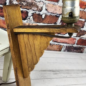 May include: A vintage wooden wall sconce with a decorative, scalloped design. The sconce features a metal light fixture and a brown electrical cord. The wood has a warm, natural finish. The sconce is mounted on a wooden base.