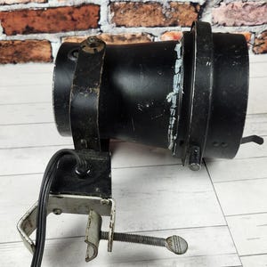 May include: A black spotlight with a clamp base, resting on a white wooden surface. The spotlight has a cylindrical body with metal bands and a small handle. A power cord extends from the base.