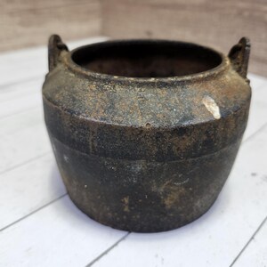 May include: A small, black cast iron pot with two handles. The pot has a rustic, aged appearance.
