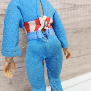 May include: A blue jumpsuit with a red and white striped belt. The jumpsuit has a zipper down the front and a seam down the back.