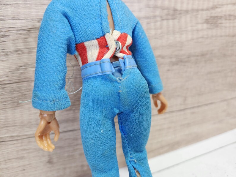 May include: A blue jumpsuit with a red and white striped belt. The jumpsuit has a zipper down the front and a seam down the back.