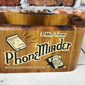 May include: Wooden Phone Minder box with a rectangular shape and rounded corners. The box has a light brown finish and features the text "Phone Minder" with illustrations of a phone and a notepad. The text "3 Min. Timer" is also visible.