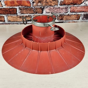 May include: A round, terracotta-colored plastic Christmas tree stand featuring a wide, ribbed base and a central cylindrical holder. The holder includes a silver-toned metal ring at the top with three adjustable metal eye bolts for securing a tree.