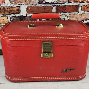 May include: A vintage red Samsonite train case with a handle and brass-colored hardware. The case has white stitching around the edges and a textured surface. The Samsonite logo is visible on the front.