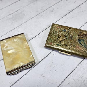 May include: Two vintage compact cases. One is gold with an intricate design of an anchor, flowers, and other nautical elements. The other is a white mother-of-pearl case with a silver trim.