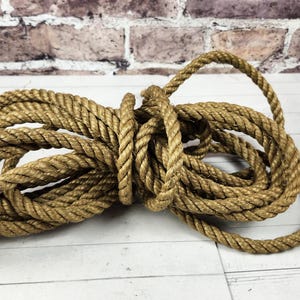 May include: Coiled natural fiber rope, tan in color, lies on a white wooden surface. The rope is thick and appears to be made of twisted strands. The background features a brick wall.