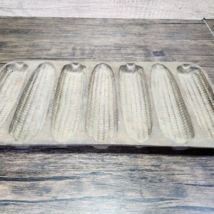 May include: A vintage cast iron cornbread pan with eight individual molds. The pan is made of a dark gray metal and has a rustic, worn look.