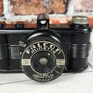May include: Vintage black Falcon Minette camera with a silver lens ring. The camera has a rectangular body with a silver winding knob and the text "FALCON MINETTE" on the lens. Made by Utility Mfg. Co. Chicago, U.S.A.