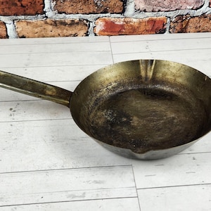 May include: A well-used, antique-style frying pan with a long handle. The pan is made of metal with a dark, aged finish. The pan is round with a slightly sloped side and a long handle with a hole at the end for hanging.