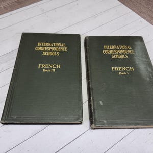 May include: Two vintage green books titled "International Correspondence Schools French" with gold lettering. One book is labeled Book III, the other Book I. The books are on a light wood surface.