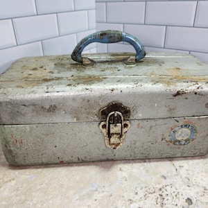 May include: Vintage metal toolbox with a blue handle and a latch closure. The toolbox has a weathered, distressed appearance with signs of rust and wear. A small sticker is visible on the side. The toolbox is rectangular and appears to be made of steel.