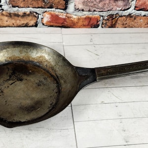 May include: A well-worn, antique-style frying pan with a long handle and a small hole at the end for hanging. The pan is made of dark metal with signs of use and wear, suggesting it's been used for cooking. The pan is on a white wooden surface.