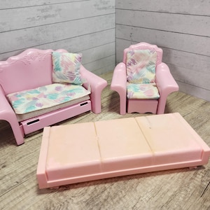 May include: A pink dollhouse furniture set featuring a sofa, armchair, and coffee table. The sofa and armchair have patterned cushions and pillows. The coffee table is rectangular. The set is made of plastic.