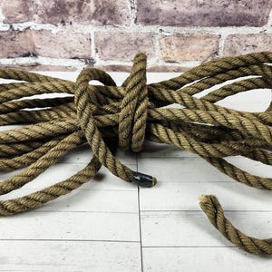 May include: Coiled brown rope with a natural, textured appearance. The rope is thick and appears to be made of natural fibers. The rope is knotted in the middle and lies on a white wooden surface.